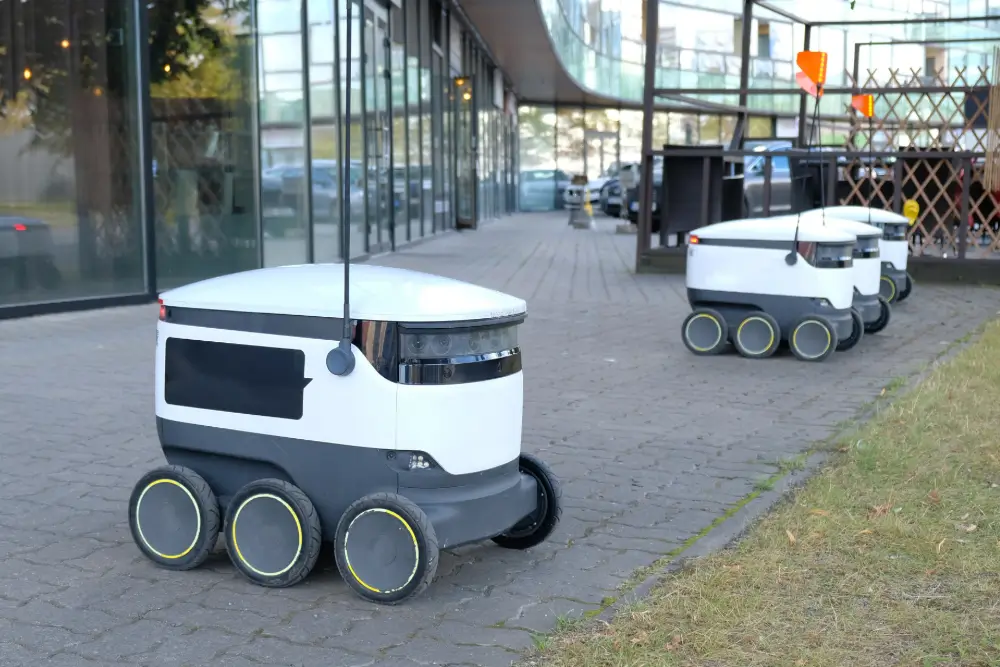 Three six-wheeled delivery robots on a paved walkway outside a glass building