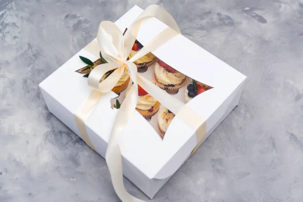 A white box of fruit-topped cupcakes tied with a cream satin ribbon, seen through its window on a grey stone surface