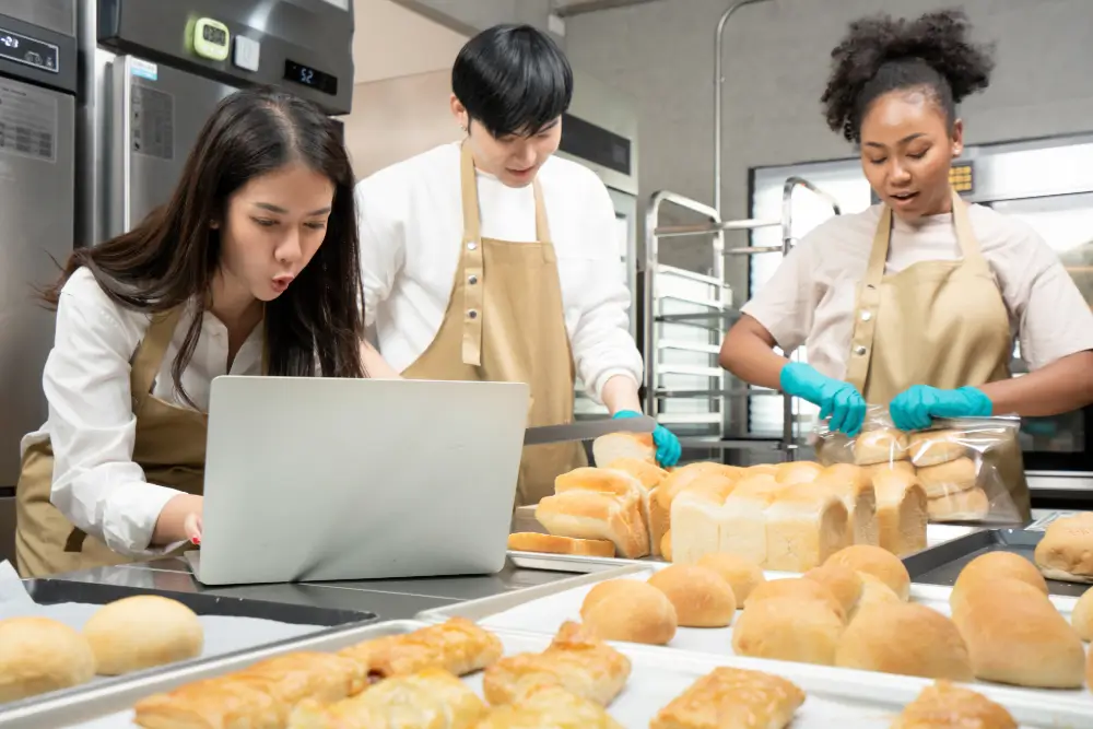 Three bakers in aprons, one checking a laptop while the others slice and bag loaves, behind trays of fresh buns and pastries