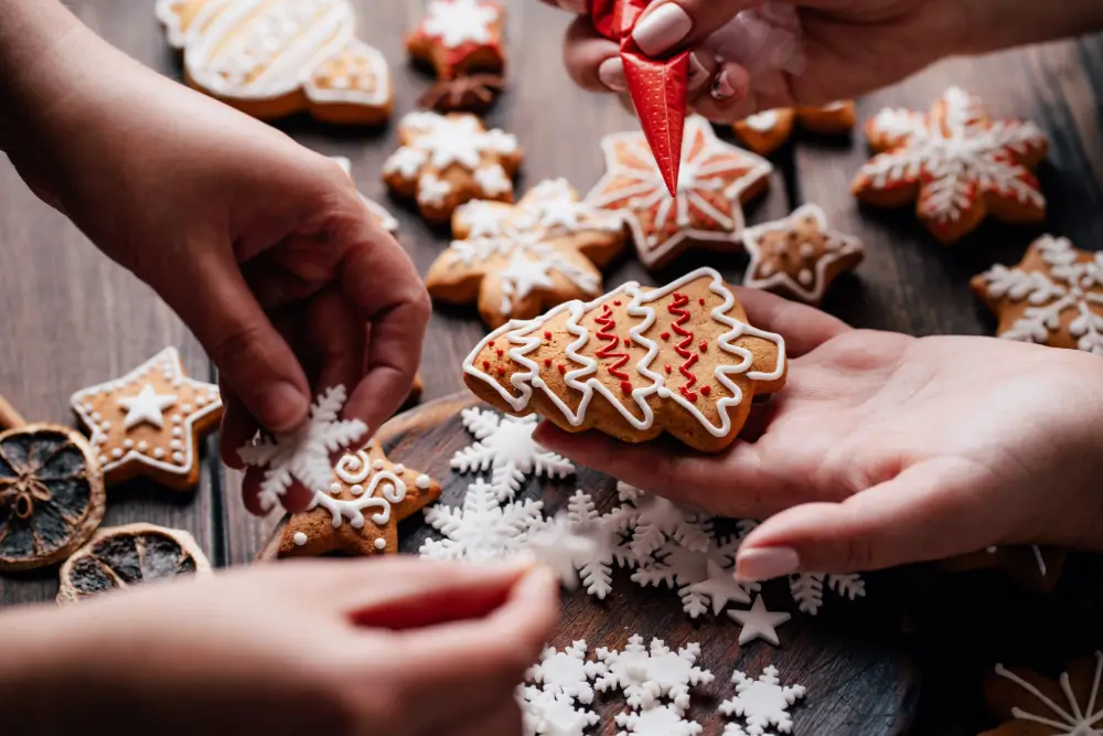 Hands piping white and red icing onto gingerbread cookies shaped like trees, stars and snowflakes