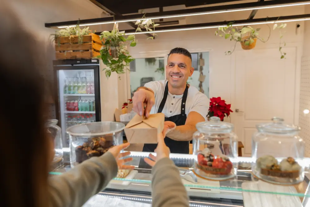A smiling baker handing a kraft paper takeout box across a pastry display case to a customer