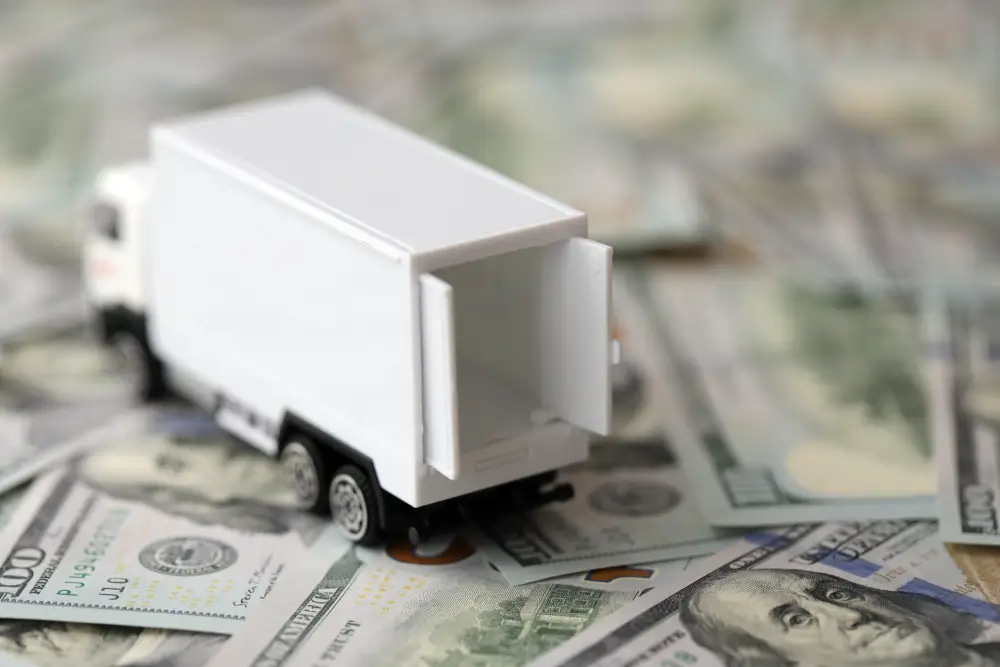 A white toy box truck with its rear doors open, parked on a spread of US hundred-dollar bills