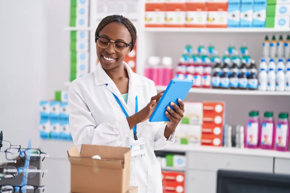 A smiling pharmacist in a white coat checking a tablet beside an open delivery box, in front of stocked pharmacy shelves