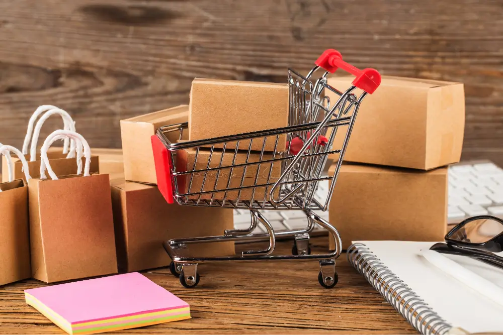 A miniature shopping cart among cartons, paper bags, a notebook and sticky notes on a wooden desk
