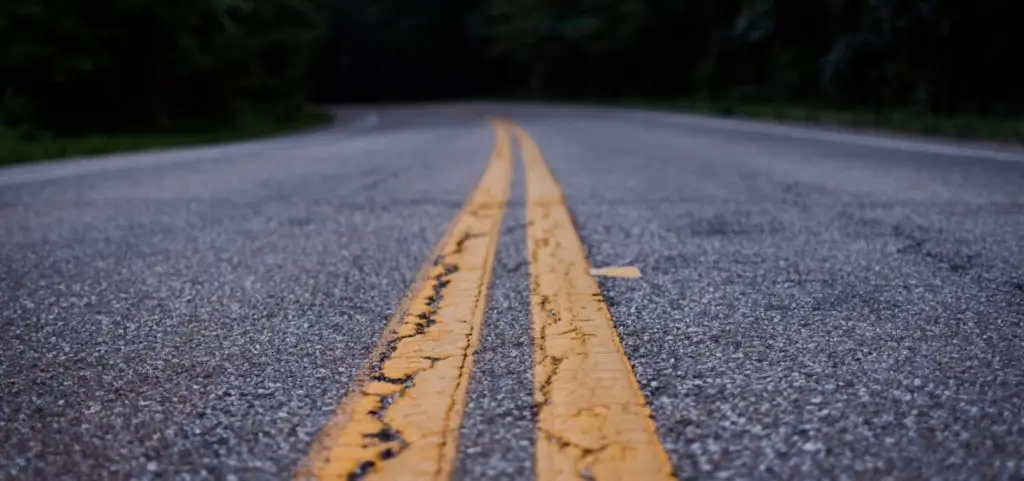 A low view along a worn asphalt road with faded double yellow centre lines curving into dark trees
