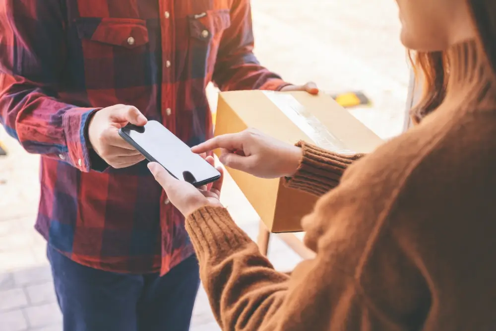A courier holding a parcel and a phone while the customer taps the screen to confirm delivery