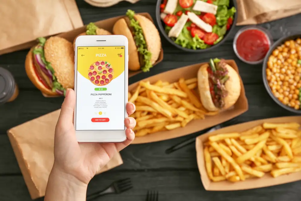 A hand holding a phone showing a pizza order screen above a table of burgers, fries and salads