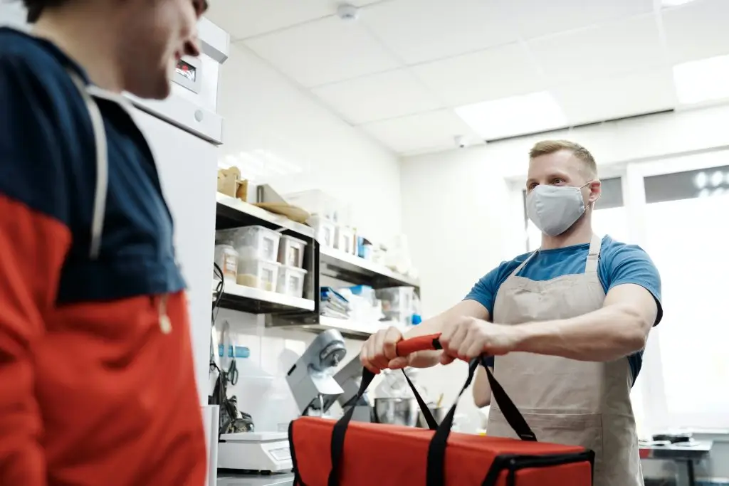 A masked cook in an apron handing a red insulated delivery bag to a courier across a bright commercial kitchen with stocked shelves