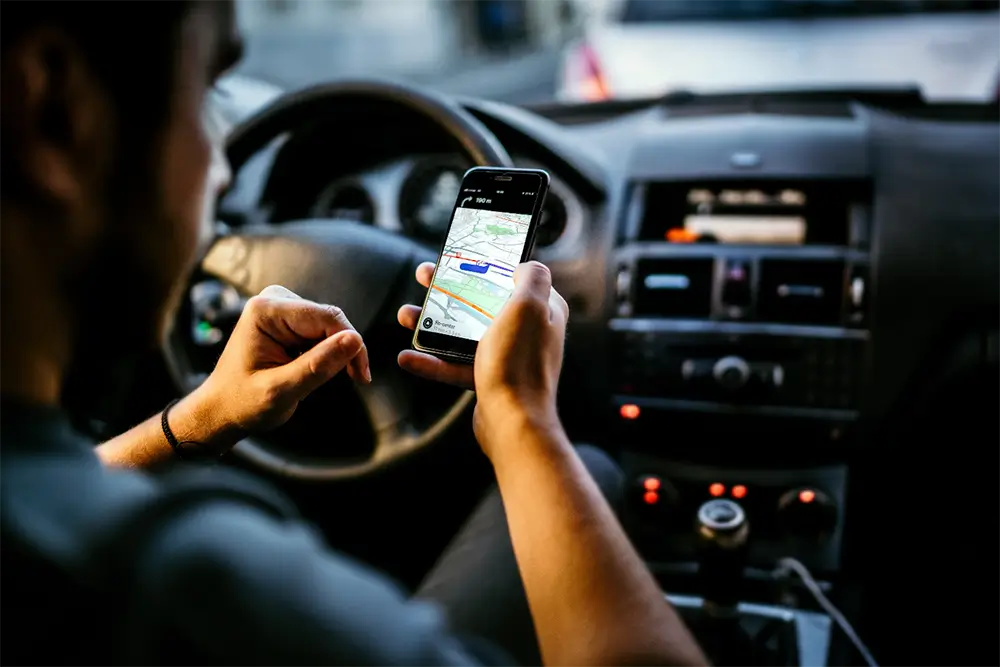A driver at the wheel of a car holding a phone showing turn-by-turn navigation on a map