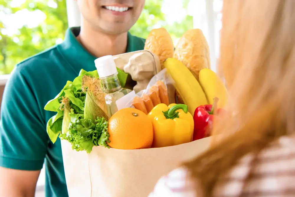 A smiling delivery worker handing over a paper bag full of groceries, with bread, bananas, peppers, lettuce and an orange, to a customer at the door