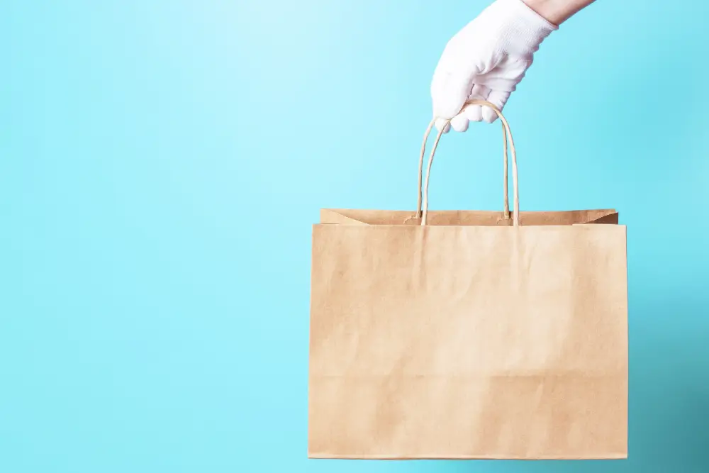 A white-gloved hand holding up a brown paper shopping bag against a light blue background