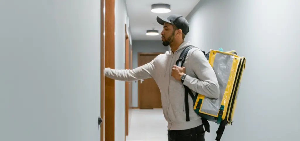 A courier with an insulated yellow and grey delivery backpack knocking on an apartment door in a hallway