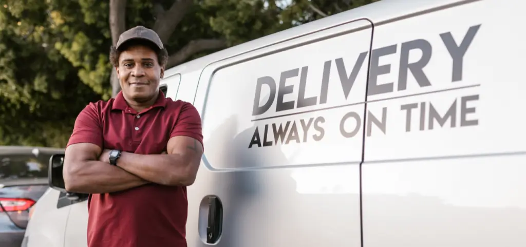 A driver standing with folded arms beside a van lettered Delivery, Always On Time