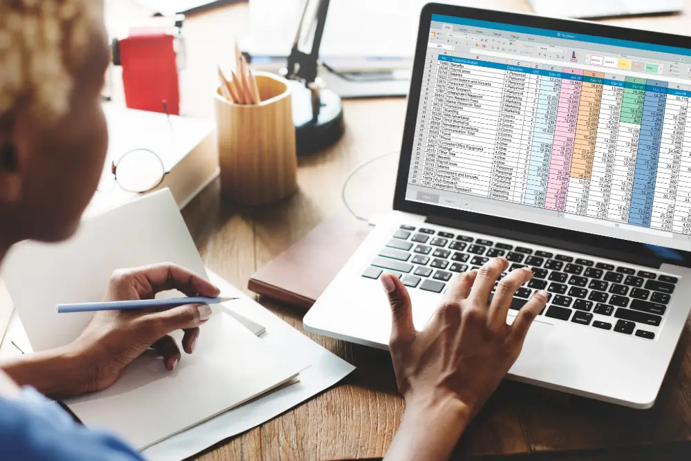 Someone working through a colour-coded spreadsheet on a laptop while making notes on a pad
