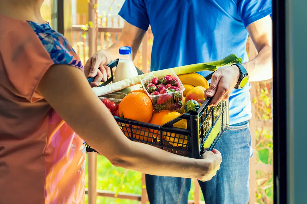 A courier handing a crate of fresh fruit, strawberries, leeks and milk to a customer at the door