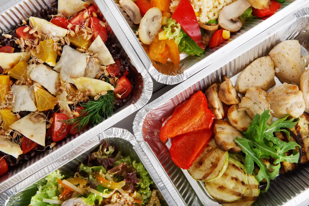 An overhead view of four foil trays of prepared meals: salads, grilled vegetables and chicken