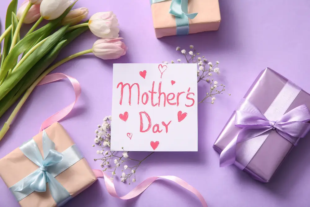A handwritten “Mother’s Day” card with red hearts on a lilac table, surrounded by pink tulips, baby’s breath and wrapped gifts