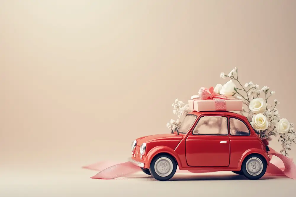 A red toy car carrying a pink gift box and white roses on its roof, trailing a pink ribbon against a beige backdrop