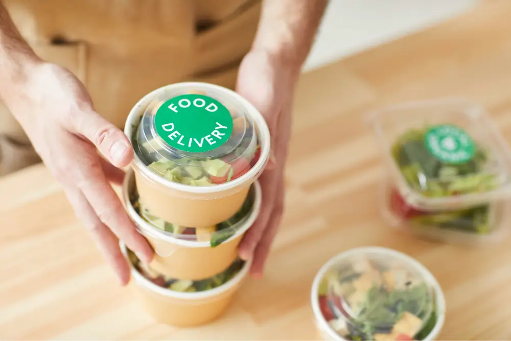 Hands lifting a stack of salad tubs sealed with green Food Delivery labels