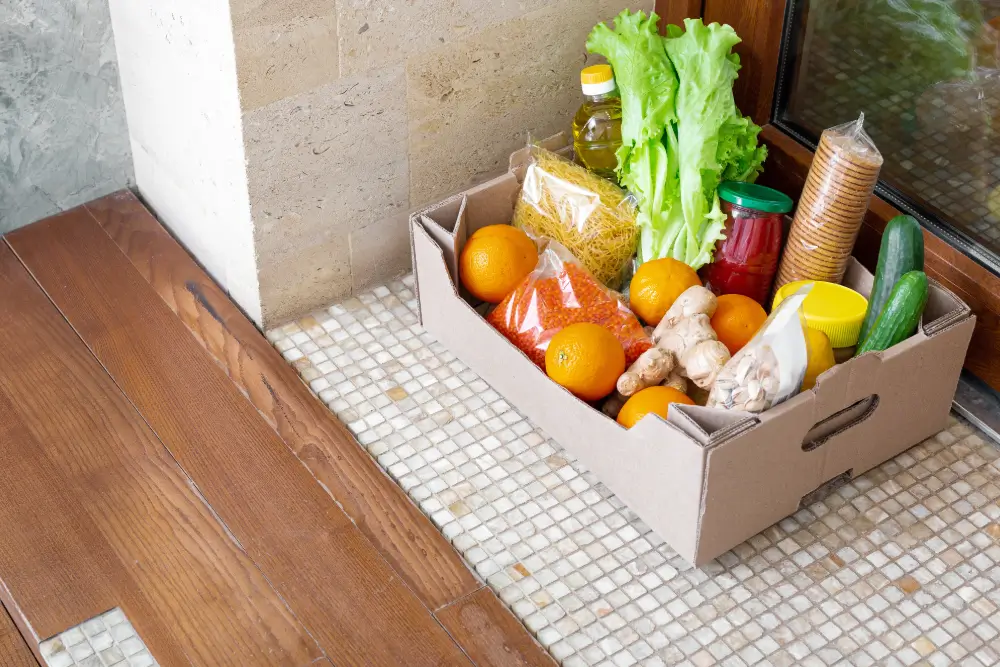 A cardboard box of groceries, including oranges, lettuce, cucumbers and ginger, left on a tiled floor by a door