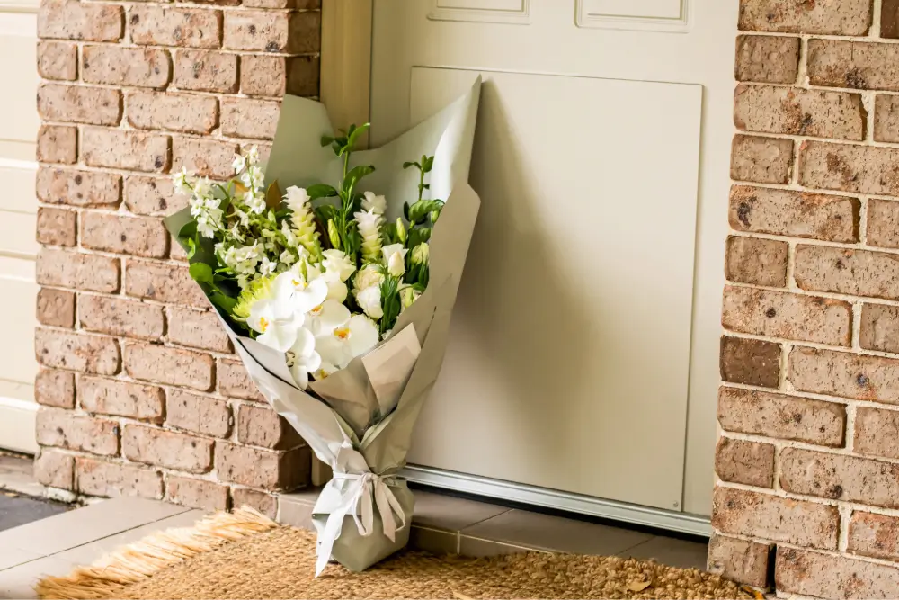 A bouquet of white orchids and lilies wrapped in grey paper, left leaning against a front door on a coir doormat between brick walls