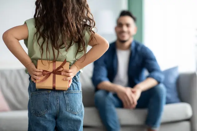A child hides a ribbon-tied gift box behind their back while a smiling adult waits on a sofa