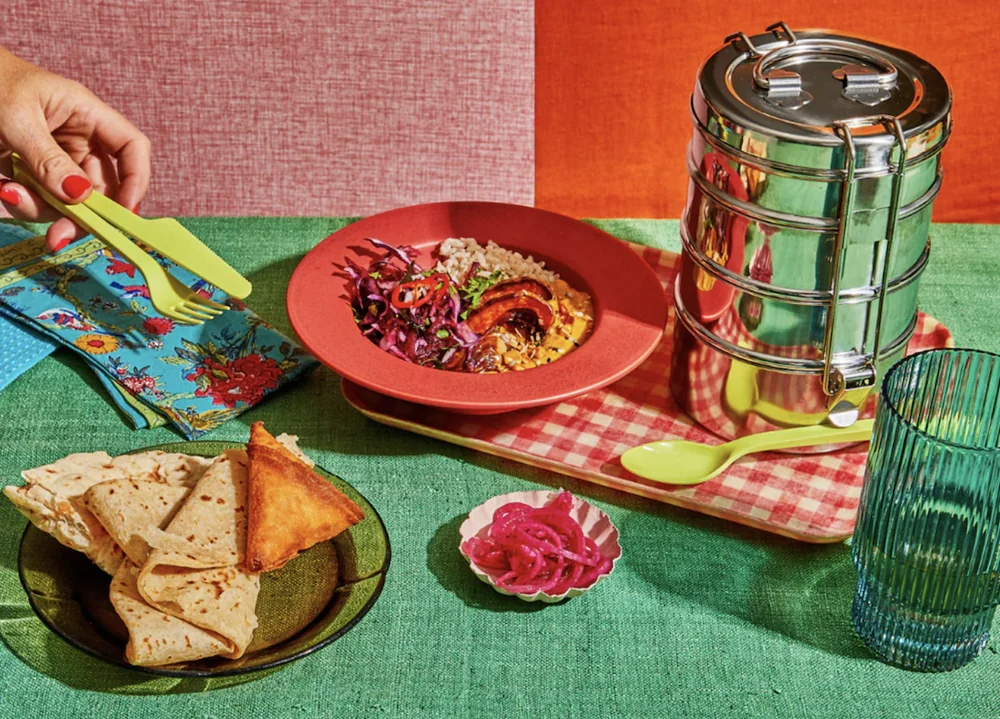 A stainless steel tiffin carrier beside a bowl of rice, roasted squash and slaw, with roti and pickled onions on a green tablecloth