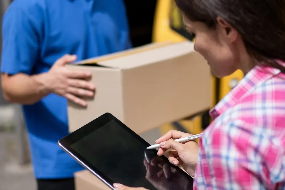 A customer signing a tablet with a stylus as a courier in a blue shirt holds out a package beside a yellow van