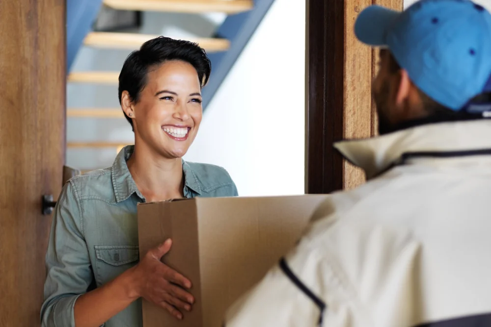 A smiling customer at the front door taking a cardboard box from a courier in a blue cap