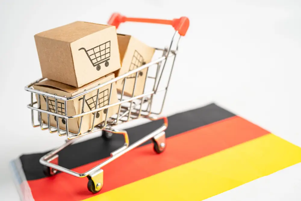 A miniature shopping cart holding cardboard boxes printed with cart icons, parked on a German flag