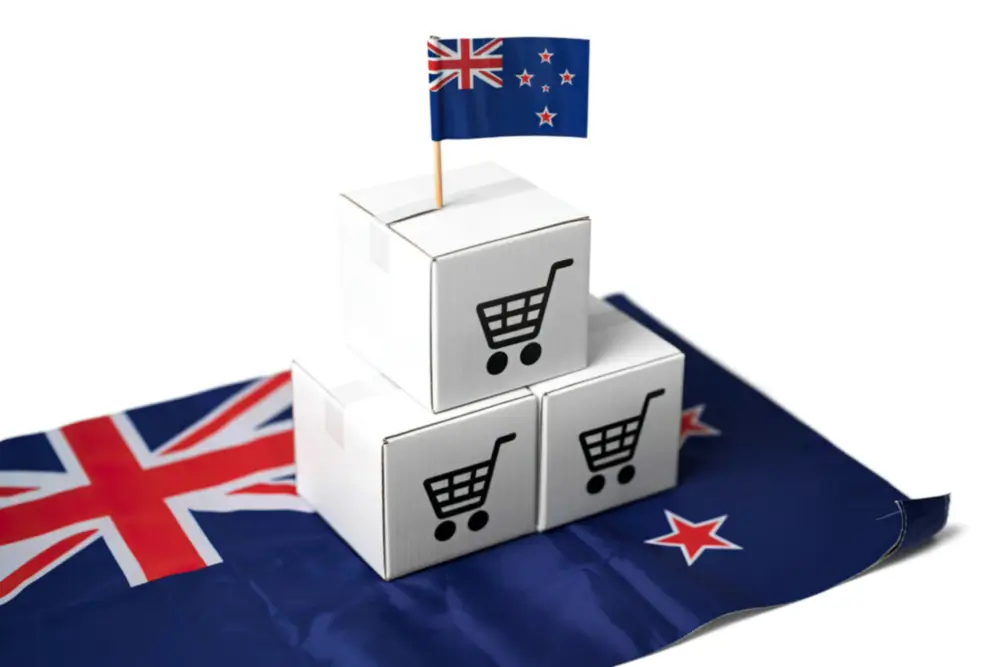Three white boxes printed with shopping cart icons, stacked on a New Zealand flag and topped with a small flag on a toothpick