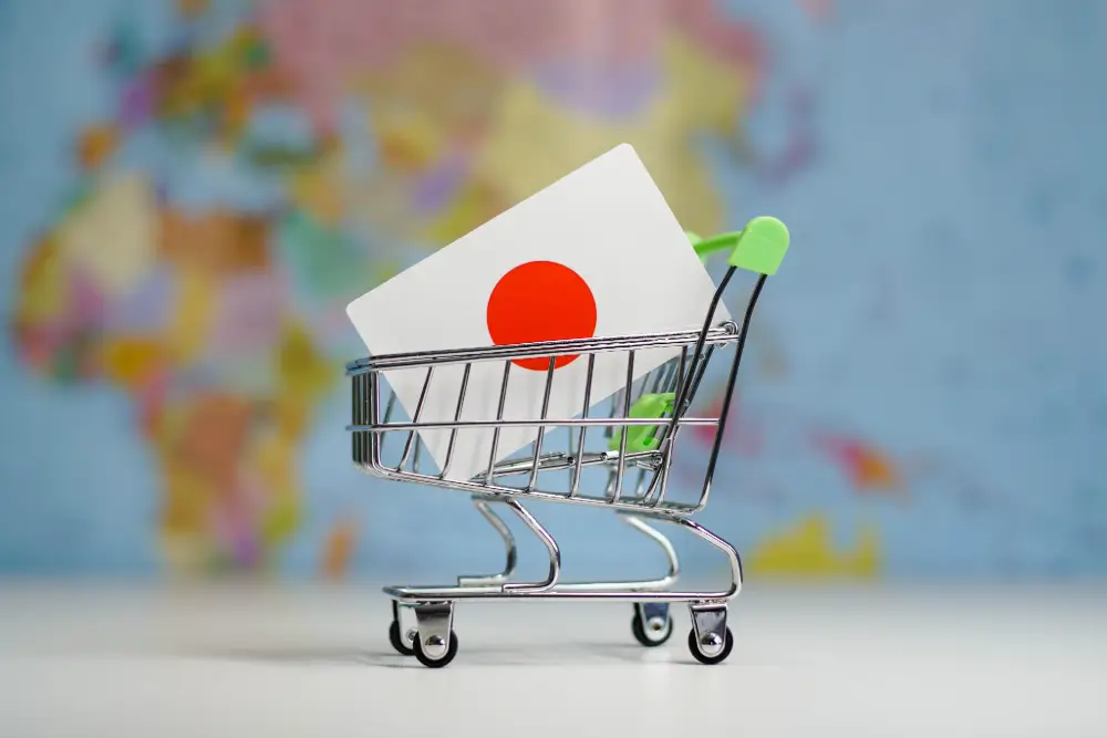 A miniature shopping cart carrying a card with the Japanese flag, in front of a blurred world map