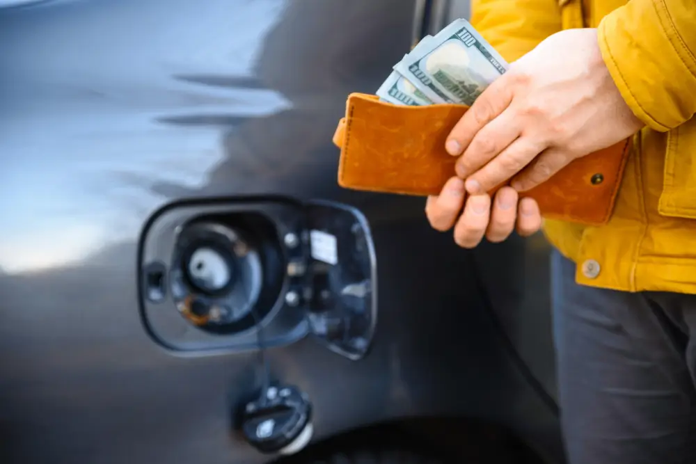 Someone in a yellow jacket pulling cash from a leather wallet beside a car’s open fuel door