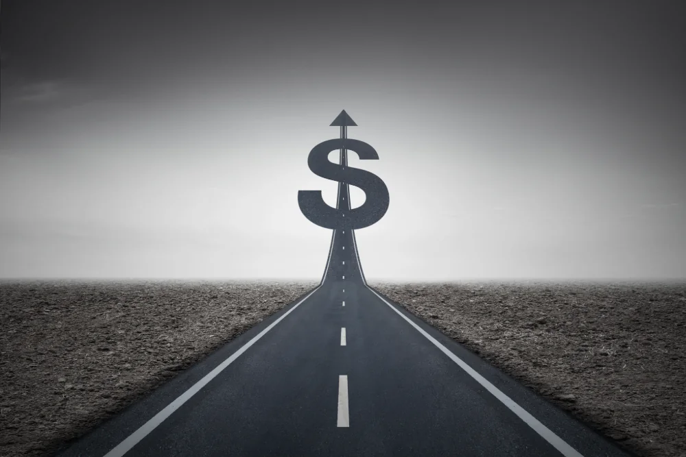 A straight asphalt highway that rises into a dollar sign topped with an arrow, against a grey sky