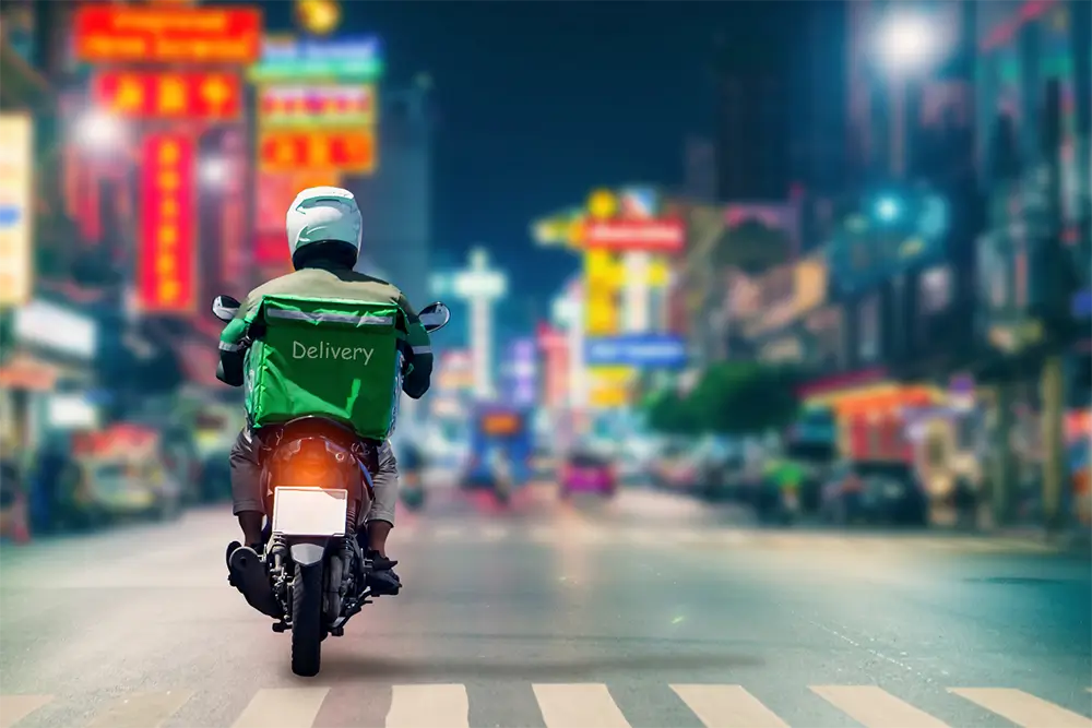 A courier on a scooter with a green Delivery box rides down a neon-lit city street at night