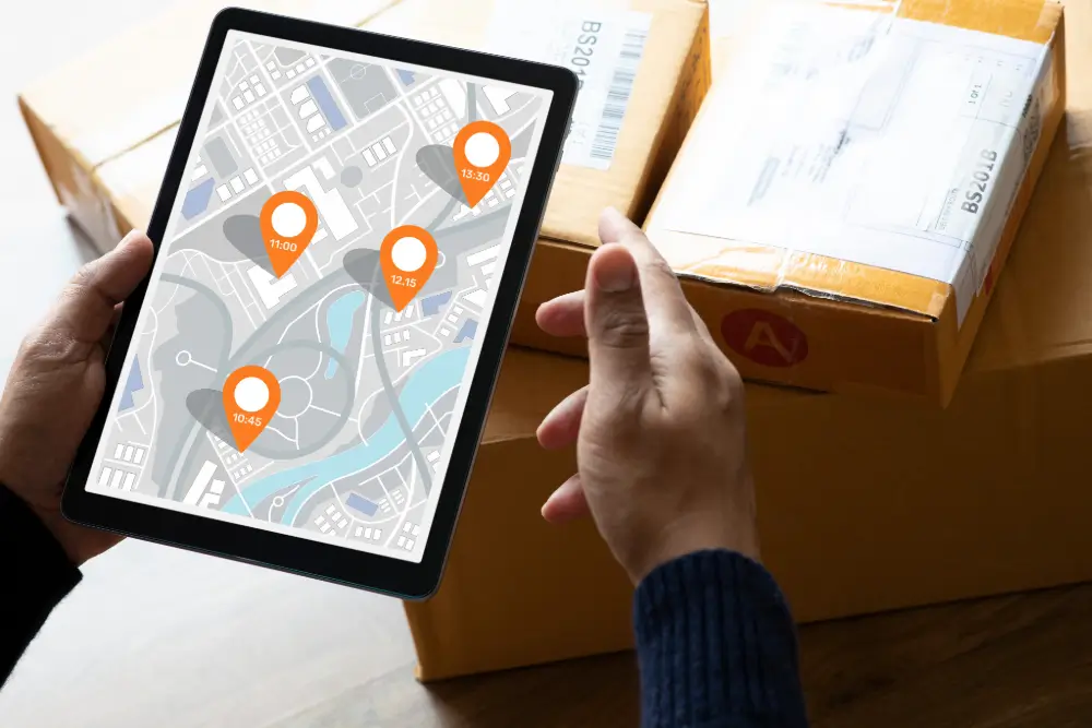 Hands holding a tablet showing a city map with four orange pins marked 10:45, 11:00, 12:15 and 13:30, in front of stacked shipping boxes