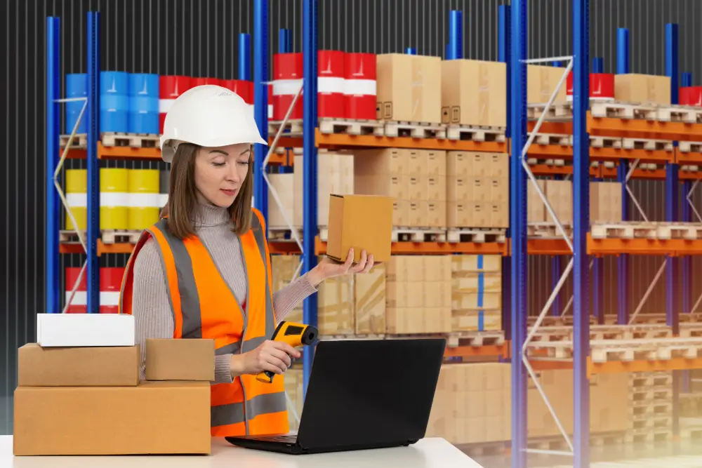 A warehouse worker in a hard hat and safety vest scanning a small box at a laptop, with stocked pallet racks behind