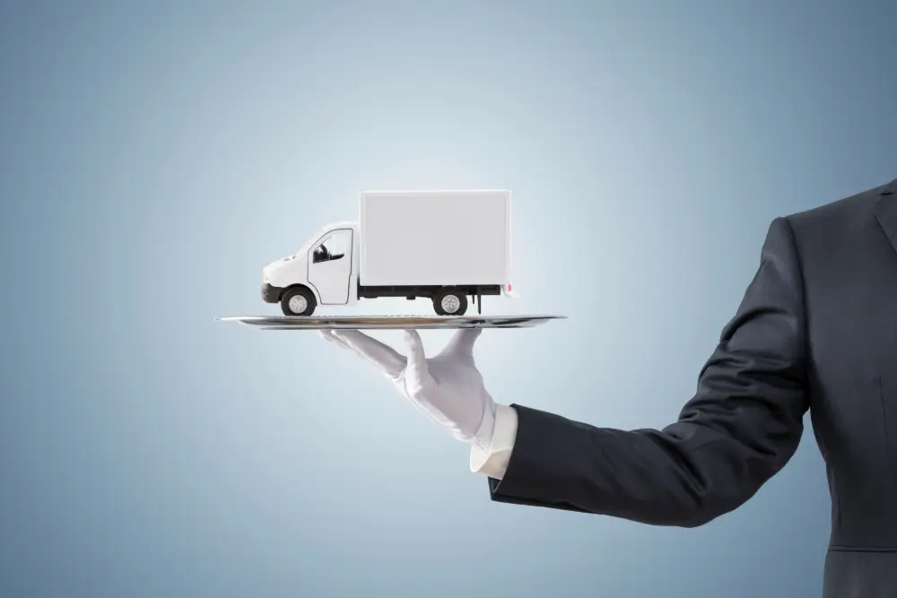 A white-gloved hand in a suit holding out a silver tray carrying a model delivery truck