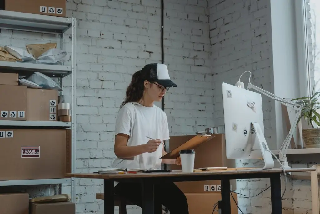 A shop worker with a clipboard at a packing desk, shelves of parcels behind her