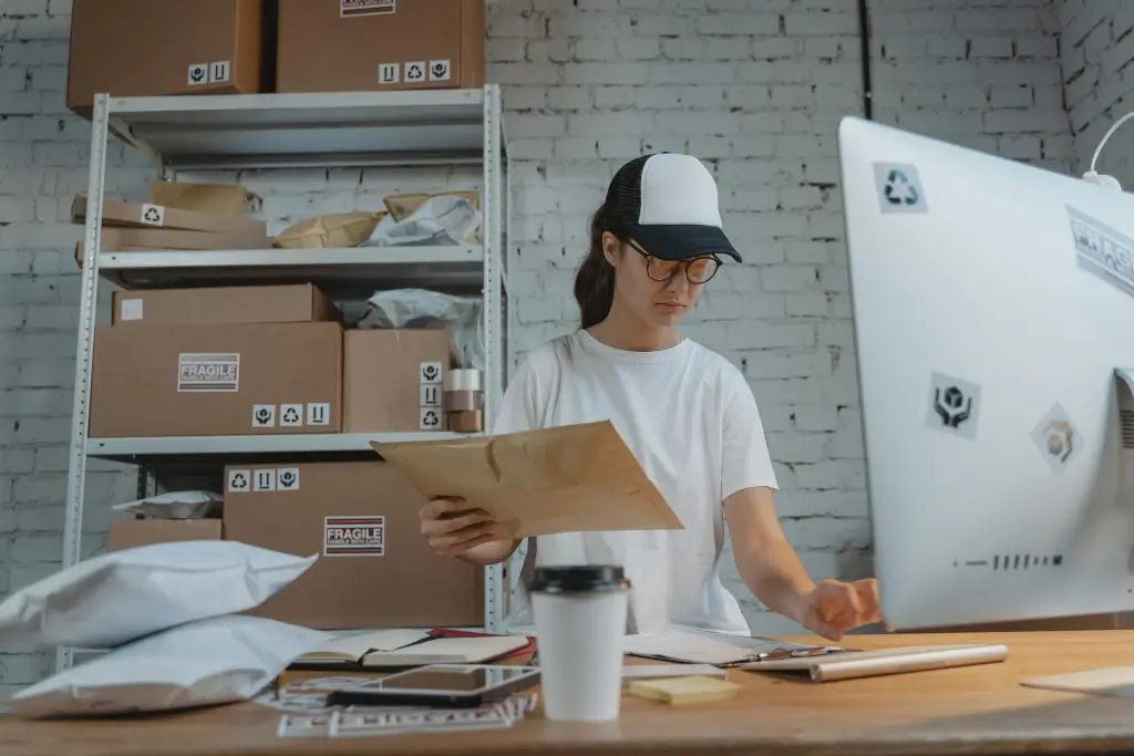 A shop worker in a cap checking a padded envelope at a packing desk, shelves of parcels behind