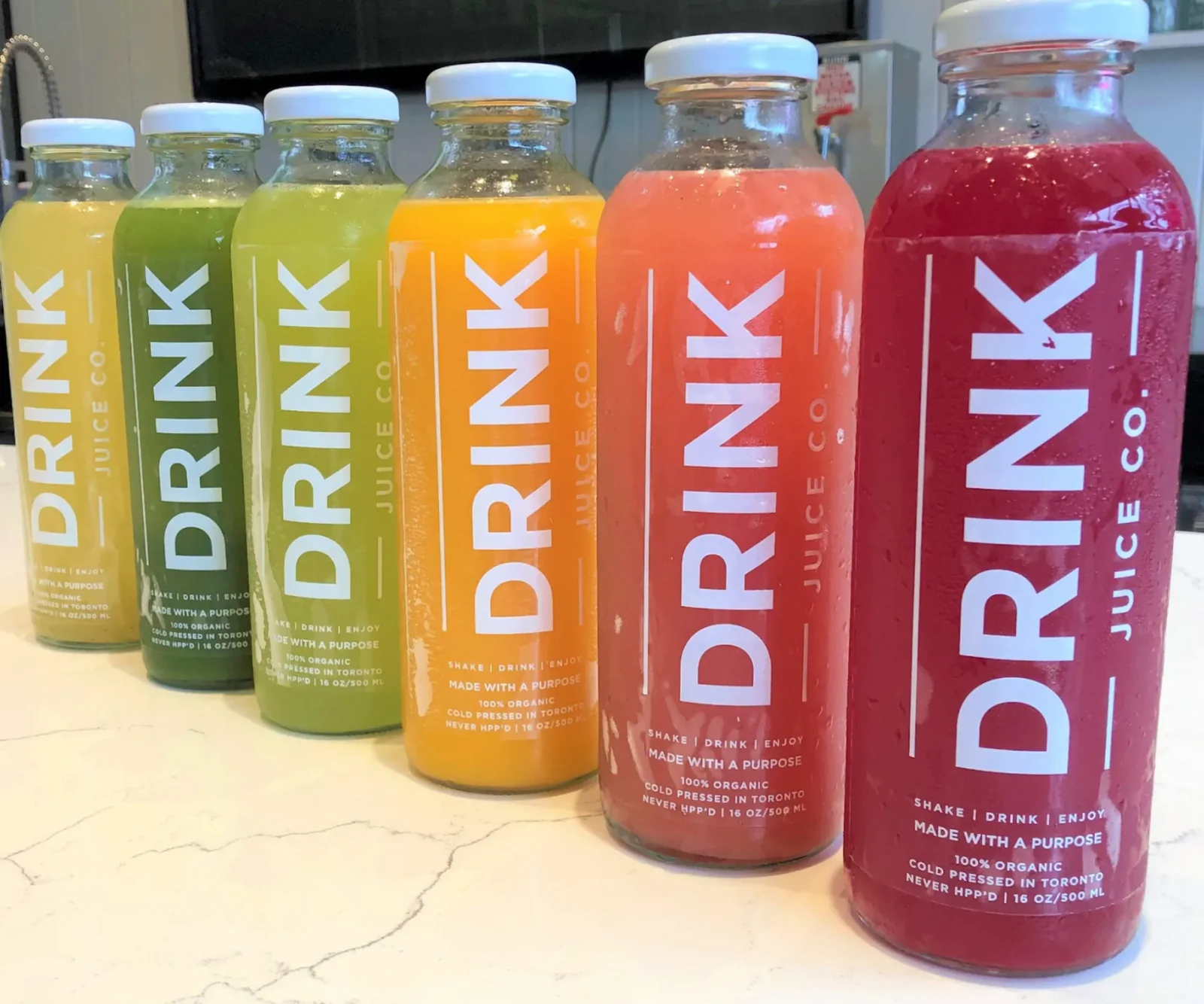 Six Drink Juice Co. bottles lined up on a counter, running from pale yellow through green and orange to deep red
