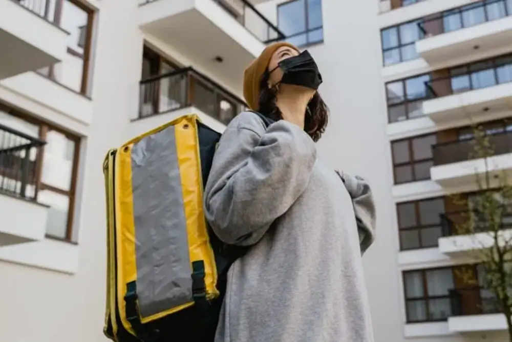 A courier in a mask and beanie with a yellow insulated backpack looking up at an apartment block