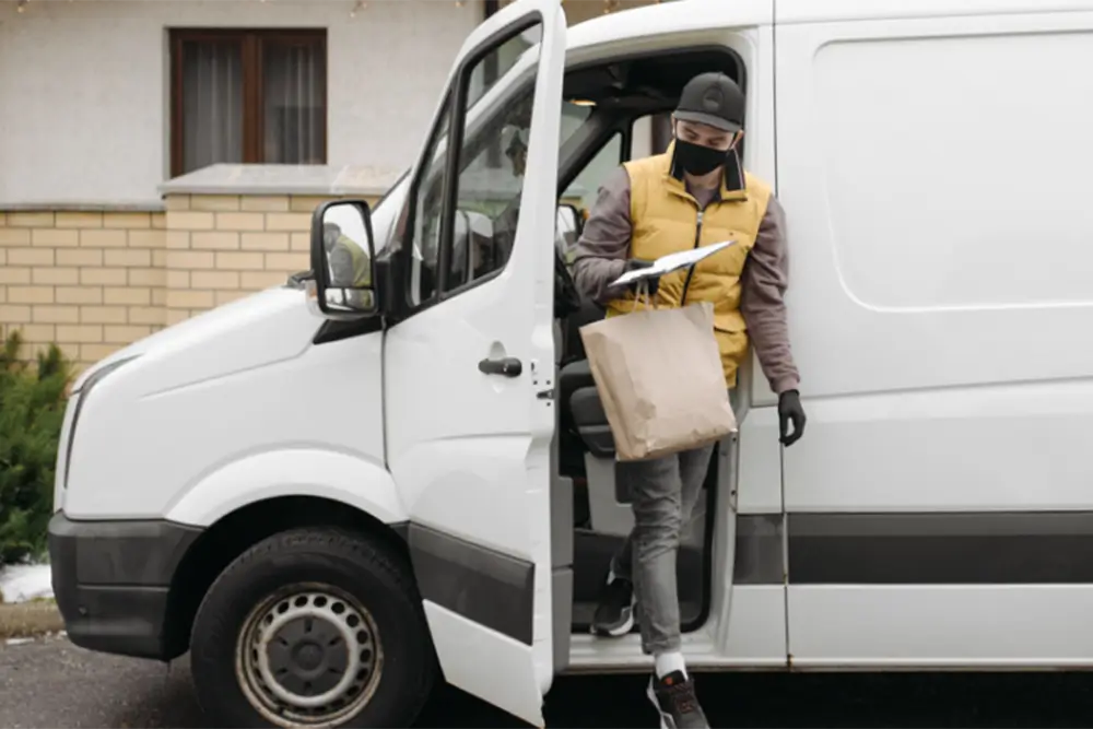 A masked courier stepping down from a van doorway with a paper bag and a clipboard