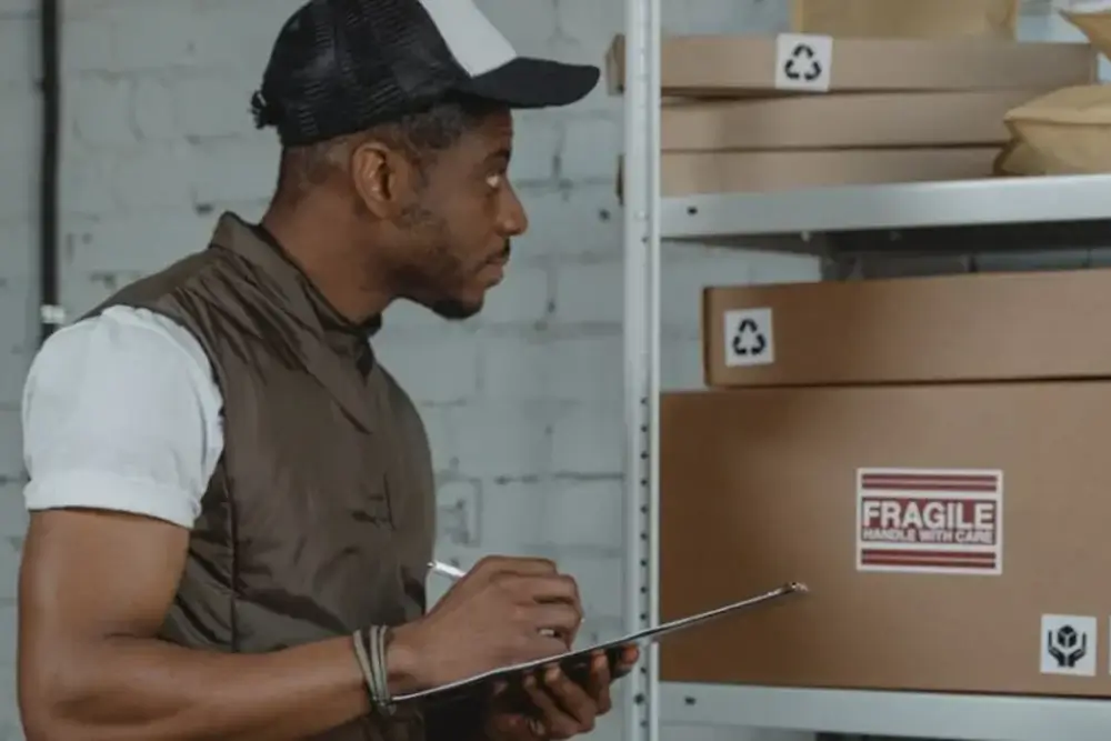 A warehouse worker with a clipboard checking cartons on a shelf, one marked Fragile, Handle With Care