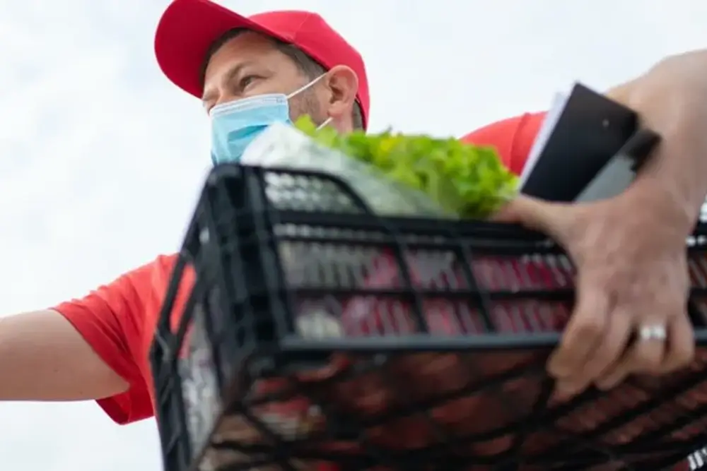 A masked courier in a red cap carrying a crate of lettuce and produce, seen from below