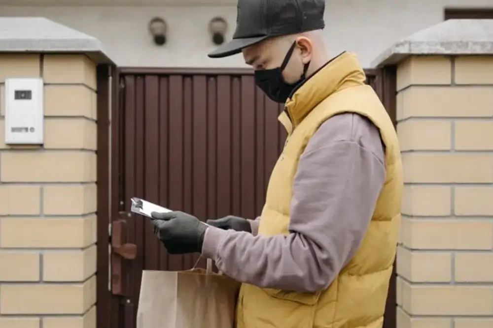 A masked courier checking a phone at a garden gate, a paper bag of groceries at his side