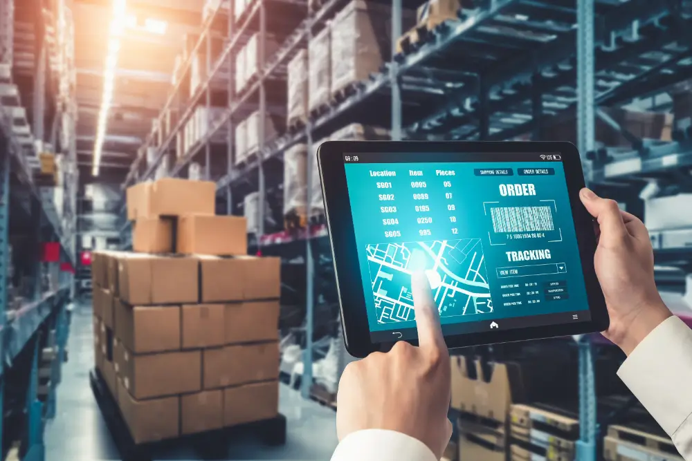 Hands using a warehouse tablet showing an order, tracking and stock list beside stacked pallets