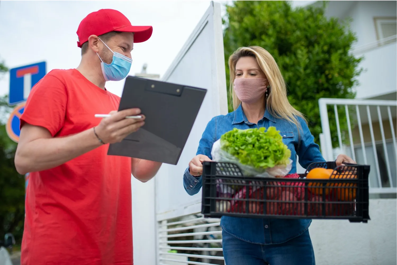 A masked delivery driver with a clipboard handing a crate of produce to a customer