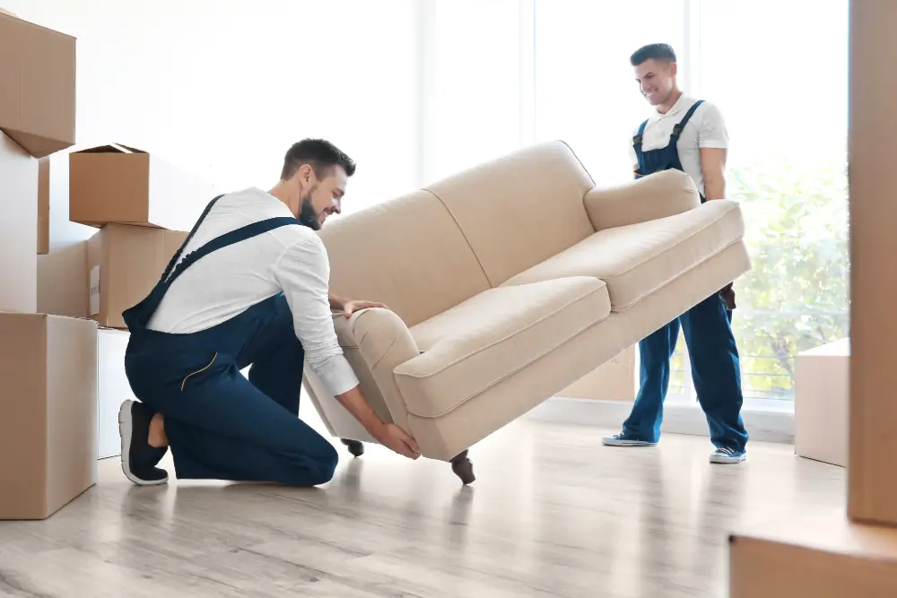 Two movers in blue overalls carrying a cream sofa into a room stacked with packing boxes