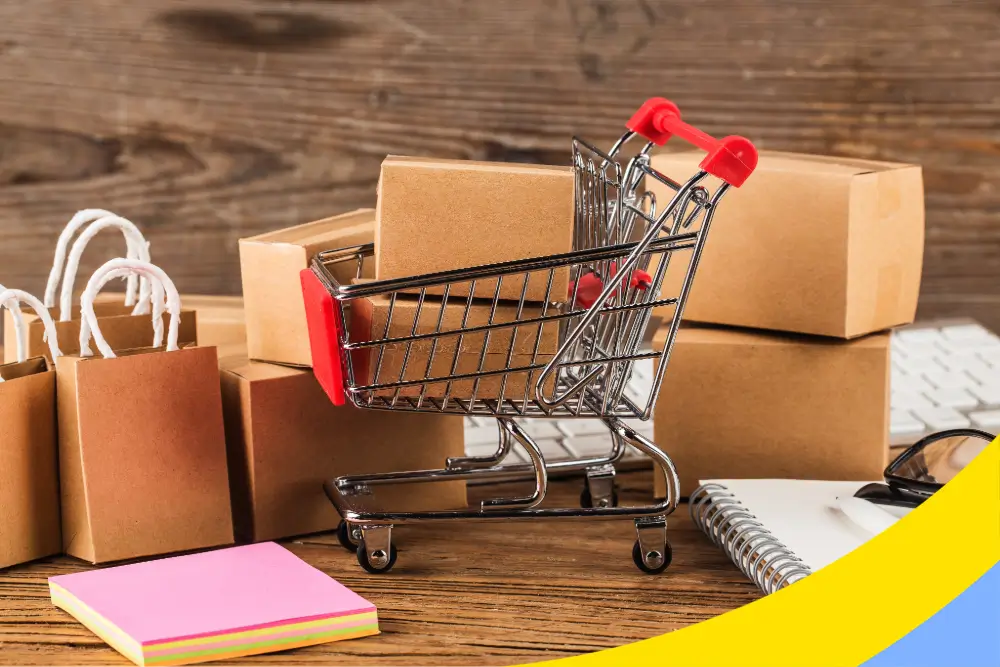 A miniature shopping cart on a desk among cartons, paper bags, a notebook and a keyboard